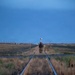 A single figure in military camouflage gear walks along railroad tracks stretching into the distance at dusk or dawn. The flat, arid landscape is flanked by dry grassland, with a small town or installation visible on the horizon, including a water tower. The low-light blue-hour conditions and receding perspective create a stark, solitary composition.