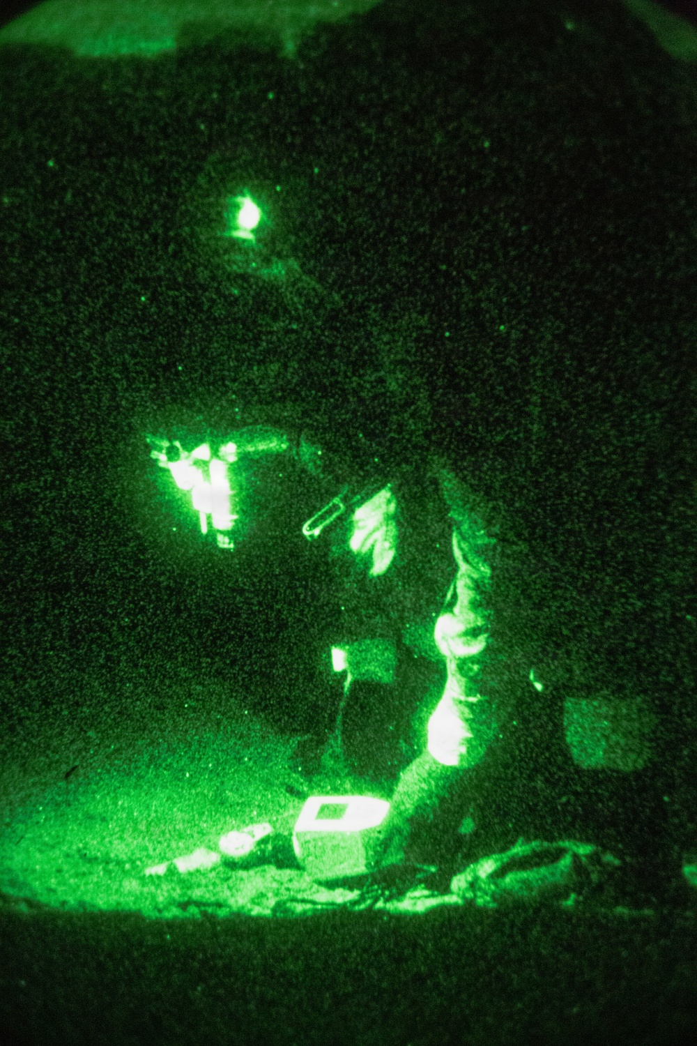 A night vision image captured through what appears to be a night vision device (NVD), showing one or more individuals in a dark outdoor environment. The green-tinted monochromatic imagery is characteristic of image intensifier night vision technology. At least one person appears to be kneeling near equipment on the ground, with bright IR light sources visible.