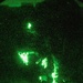 A night vision image captured through what appears to be a night vision device (NVD), showing one or more individuals in a dark outdoor environment. The green-tinted monochromatic imagery is characteristic of image intensifier night vision technology. At least one person appears to be kneeling near equipment on the ground, with bright IR light sources visible.