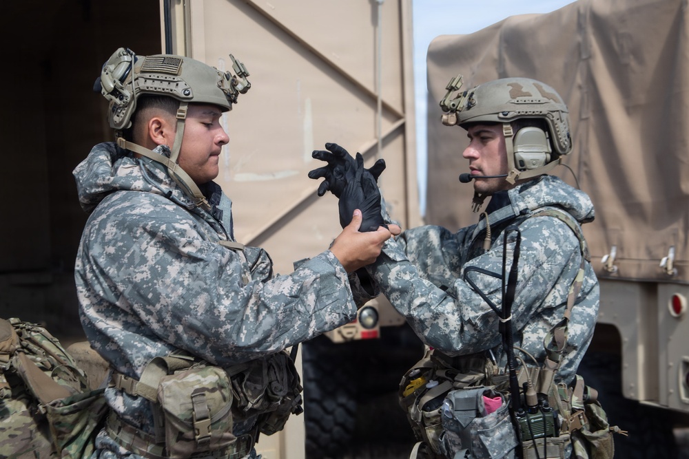 Two military personnel in full combat gear are interacting near a military vehicle, with one soldier appearing to put on or adjust a black tactical glove. Both wear digital camouflage uniforms (ACU pattern) and tactical helmets with accessories. The setting appears to be a staging or operational area with military trucks visible in the background.