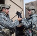 Two military personnel in full combat gear are interacting near a military vehicle, with one soldier appearing to put on or adjust a black tactical glove. Both wear digital camouflage uniforms (ACU pattern) and tactical helmets with accessories. The setting appears to be a staging or operational area with military trucks visible in the background.
