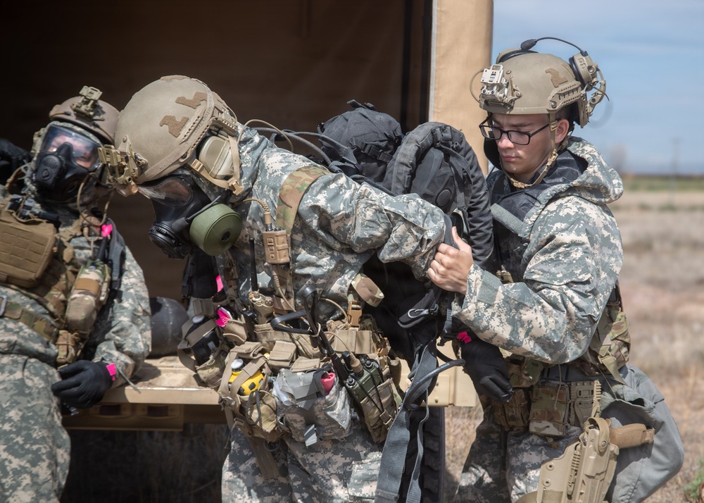 Military personnel in full combat gear are engaged in what appears to be a field training exercise or operational activity near a military vehicle or tent structure. At least three soldiers are visible, with two wearing gas masks and tactical helmets, while one soldier wearing glasses assists with handling a large backpack or equipment bag. The arid, open terrain and bright daylight conditions suggest a desert or semi-arid training environment.