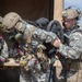 Military personnel in full combat gear are engaged in what appears to be a field training exercise or operational activity near a military vehicle or tent structure. At least three soldiers are visible, with two wearing gas masks and tactical helmets, while one soldier wearing glasses assists with handling a large backpack or equipment bag. The arid, open terrain and bright daylight conditions suggest a desert or semi-arid training environment.
