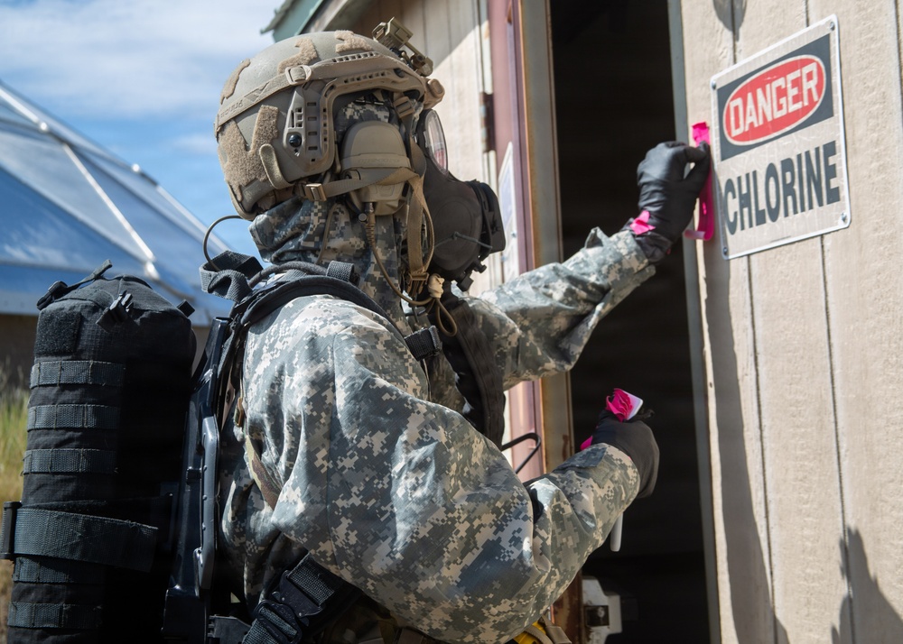 A military personnel wearing full CBRN (Chemical, Biological, Radiological, and Nuclear) protective gear is approaching or marking a building with a 'DANGER CHLORINE' warning sign. The soldier is equipped with a gas mask, tactical helmet, ACU (Army Combat Uniform) camouflage, black gloves, and what appears to be a self-contained breathing apparatus (SCBA) on their back. This appears to be a CBRN training exercise or hazmat response scenario.