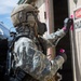A military personnel wearing full CBRN (Chemical, Biological, Radiological, and Nuclear) protective gear is approaching or marking a building with a 'DANGER CHLORINE' warning sign. The soldier is equipped with a gas mask, tactical helmet, ACU (Army Combat Uniform) camouflage, black gloves, and what appears to be a self-contained breathing apparatus (SCBA) on their back. This appears to be a CBRN training exercise or hazmat response scenario.