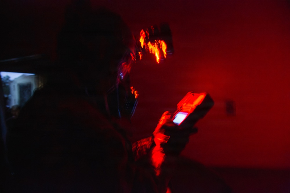 A person is shown in a dark environment dominated by intense red lighting, holding what appears to be a handheld electronic device with a small illuminated screen. The scene is heavily motion-blurred, particularly around the subject's head area, suggesting camera movement during a long exposure. The red ambient light and dark surroundings create a dramatic, high-contrast atmosphere.