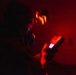 A person is shown in a dark environment dominated by intense red lighting, holding what appears to be a handheld electronic device with a small illuminated screen. The scene is heavily motion-blurred, particularly around the subject's head area, suggesting camera movement during a long exposure. The red ambient light and dark surroundings create a dramatic, high-contrast atmosphere.