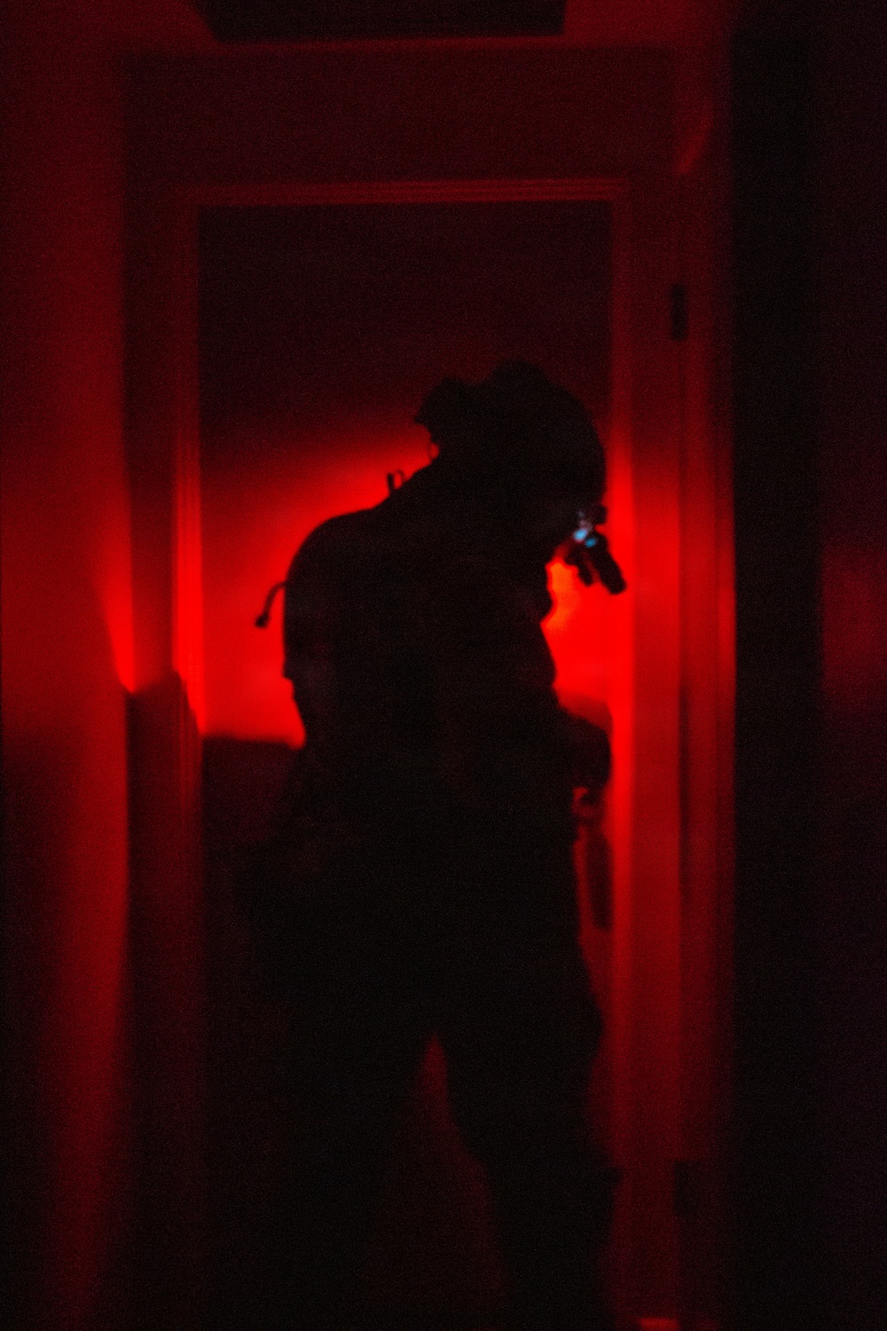 A silhouetted figure stands in a doorway or corridor illuminated by intense red lighting. The figure appears to be equipped with tactical gear including a helmet and is holding what appears to be a weapon or flashlight. The extreme low-light, red-lit environment creates a high-contrast, dramatic composition.