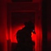 A silhouetted figure stands in a doorway or corridor illuminated by intense red lighting. The figure appears to be equipped with tactical gear including a helmet and is holding what appears to be a weapon or flashlight. The extreme low-light, red-lit environment creates a high-contrast, dramatic composition.