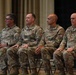 Four U.S. Army soldiers in Operational Camouflage Pattern (OCP) uniforms are seated in folding chairs on what appears to be a stage or auditorium setting. They appear to be attending a formal military ceremony or event, sitting attentively in a row. Name tapes are partially visible on at least three individuals, with readable names including 'FOUST' and 'BATES'.