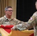 Two U.S. Army soldiers in OCP (Operational Camouflage Pattern) uniforms are participating in a formal military ceremony, appearing to unfurl or case a unit guidon or colors. The soldier facing the camera is identified by his name tape as 'BRITTAIN' and wears a Colonel's eagle insignia along with a 3rd Infantry Division patch. The ceremony appears to be a change of command or activation/deactivation event taking place indoors.