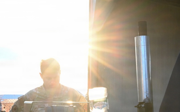 A person in military or tactical clothing is working behind a piece of heavy equipment or machinery, likely outdoors. The image is heavily backlit by direct sunlight, creating strong lens flare and silhouetting effects. A second person in dark clothing is partially visible on the right edge of the frame.