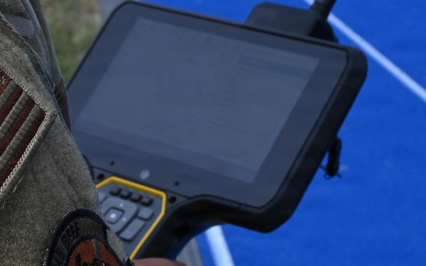 A U.S. military personnel member in OCP (Operational Camouflage Pattern) uniform is holding a ruggedized tablet or remote control device with a yellow and black housing. The individual displays a U.S. flag patch and a unit patch featuring a tiger emblem on their left sleeve. The background shows a blue surface, likely an athletic court or tarmac area with white line markings.