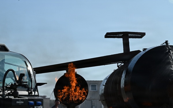 The image depicts a firefighting training exercise at what appears to be a military or airport fire training facility. Firefighters in full turnout gear are positioned near an Oshkosh Striker ARFF (Aircraft Rescue and Fire Fighting) vehicle alongside a simulated aircraft fuselage trainer engulfed in controlled flames. The training prop replicates a helicopter or aircraft fuselage with rotor components to simulate realistic crash fire scenarios.