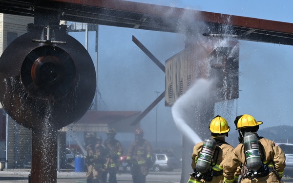 A group of firefighters in full protective gear and SCBA (self-contained breathing apparatus) are conducting what appears to be a firefighting training exercise at an industrial or military training facility. Three firefighters in the foreground are operating a yellow hose, directing a water stream toward a structure, while additional firefighters are visible in the background. Small flames are visible in the lower-left corner near a drain grate, suggesting a controlled burn scenario.