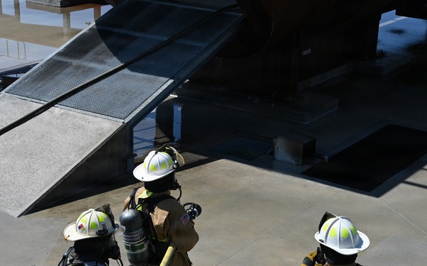 Three firefighters in full protective gear are positioned near what appears to be a training structure or ramp, holding a fire hose. They are equipped with SCBA (Self-Contained Breathing Apparatus) tanks and wearing standard turnout gear with reflective striping and white helmets. The scene appears to be a firefighting training exercise conducted on a concrete pad.