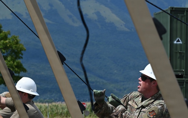 Military personnel in camouflage uniforms and white hard hats are assembling or erecting a large frame structure, likely a tent or temporary shelter. The scene appears to be an outdoor military installation or exercise area, with green storage containers and mountainous terrain visible in the background. One service member appears to be giving verbal instructions or commands while handling cables or ropes.