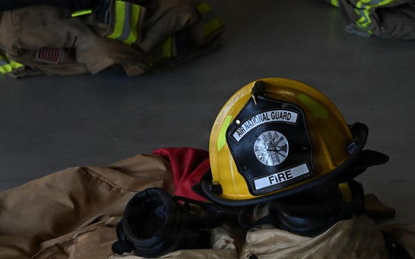 The image shows firefighting turnout gear laid out on a concrete floor, including a yellow helmet marked 'Air National Guard FIRE' resting on top of bunker pants and a jacket. The equipment appears to be staged in a ready position, likely for a quick-don drill or inspection. Additional sets of gear are visible in the blurred background.