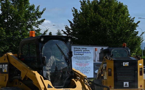 Two yellow Caterpillar (CAT) construction machines are operating at an active construction or paving site. The foreground machine is a CAT 279D compact track loader fitted with a cylindrical roller attachment, likely used for compacting ground surface. A billboard advertising a 'FreshStart' smoking cessation program is visible in the background, along with trees, mountains, and a fenced perimeter.