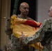 Two U.S. Army soldiers in OCP (Operational Camouflage Pattern) uniforms are participating in a formal military ceremony involving the unfurling or casing of a unit guidon or colors. The flag appears to be yellow/gold and red with gold fringe, consistent with a U.S. Army unit flag. The ceremony is taking place indoors, likely in an auditorium or assembly hall with other soldiers seated in the background.