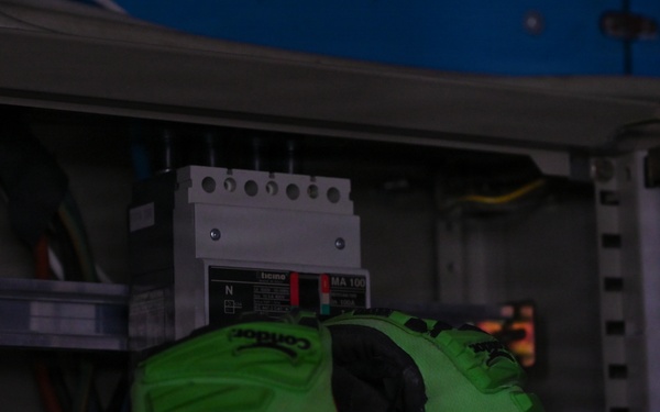 A technician wearing bright green and black Condor brand work gloves is working inside an electrical panel or distribution box. The panel contains a circuit breaker or disconnect switch labeled 'MA 10' with visible red indicators, along with multiple colored wires. The image is taken in low-light conditions, making some details difficult to discern.