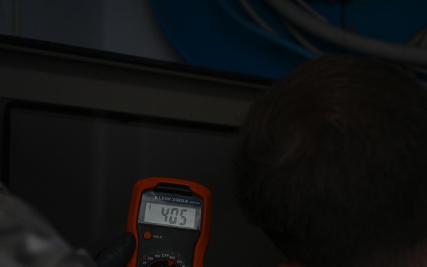 Two individuals in military-style camouflage uniforms are examining a Klein Tools digital multimeter in a dimly lit environment. The multimeter display reads '405', and the person holding it is wearing black gloves. They appear to be conducting an electrical measurement or diagnostic task near what looks like an electrical panel or equipment enclosure.
