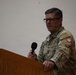 A U.S. Army officer in Operational Camouflage Pattern (OCP) uniform stands at a wooden podium holding a handheld microphone, apparently delivering a speech or address. His name tape reads 'BRITAIN' (or similar) and he wears a colonel's eagle rank insignia along with a unit patch on his right shoulder. The setting appears to be an indoor venue or gymnasium.