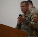 A U.S. Army soldier is speaking at a podium while holding a microphone, appearing to address an audience. He is wearing MultiCam (OCP) camouflage uniform with a '1st Armored Division' (Old Ironsides) patch visible on his left shoulder. A second soldier is partially visible in the background.