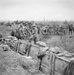 A black-and-white archival photograph depicting a World War I military scene with soldiers occupying a trench in the foreground while cavalry, horse-drawn wagons, and infantry advance across an open field in the background. The soldiers in the trench are wearing British-style Brodie helmets, suggesting this is a British or Commonwealth unit. The scene conveys active military movement, likely during an offensive operation on the Western Front.