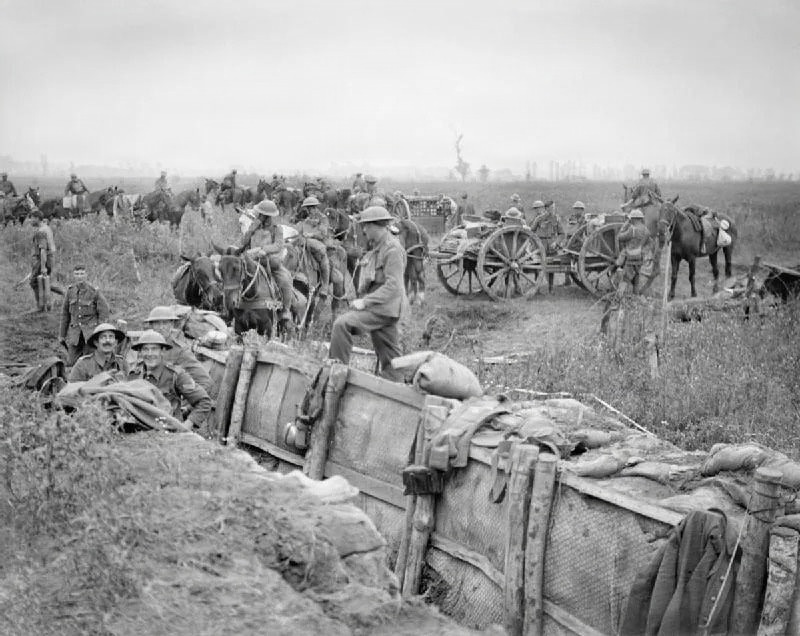 A black-and-white archival photograph depicting a World War I military scene with soldiers occupying a trench in the foreground while cavalry, horse-drawn wagons, and infantry advance across an open field in the background. The soldiers in the trench are wearing British-style Brodie helmets, suggesting this is a British or Commonwealth unit. The scene conveys active military movement, likely during an offensive operation on the Western Front.