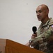 A U.S. Army Colonel is standing at a wooden podium and speaking into a handheld microphone. He is wearing the Operational Camouflage Pattern (OCP) uniform with visible U.S. Army tape, a unit patch on the right shoulder, and a Colonel's eagle rank insignia. The setting appears to be an indoor briefing or ceremony.