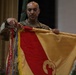 A U.S. Army soldier in OCP (Operational Camouflage Pattern) uniform is handling a military guidon or organizational flag during what appears to be a formal military ceremony. The flag is gold and red with a distinctive unit insignia visible, and multiple campaign streamers are attached to the staff. The event appears to be taking place indoors in a ceremonial hall with other military personnel visible in the background.