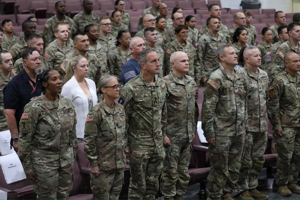 A large group of U.S. Army soldiers in OCP (Operational Camouflage Pattern) uniforms are standing at attention in what appears to be an auditorium or ceremony hall. The formation includes both male and female soldiers of various ranks, with a few civilians visible in the background. This appears to be a formal military ceremony or official event.
