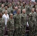 A large group of U.S. Army soldiers in OCP (Operational Camouflage Pattern) uniforms are standing at attention in what appears to be an auditorium or ceremony hall. The formation includes both male and female soldiers of various ranks, with a few civilians visible in the background. This appears to be a formal military ceremony or official event.