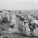 A black-and-white archival photograph showing a column of soldiers marching along a makeshift wooden plank road through a heavily devastated battlefield landscape. The terrain is littered with destroyed wagons, debris, and churned earth consistent with heavy artillery bombardment. The IWM (Imperial War Museum) watermark suggests this is a World War I era photograph.