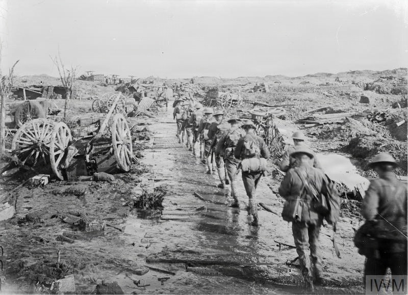 A black-and-white archival photograph showing a column of soldiers marching along a makeshift wooden plank road through a heavily devastated battlefield landscape. The terrain is littered with destroyed wagons, debris, and churned earth consistent with heavy artillery bombardment. The IWM (Imperial War Museum) watermark suggests this is a World War I era photograph.