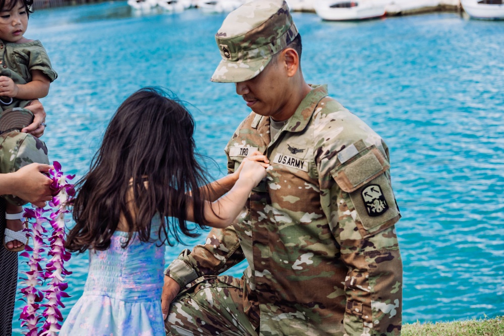 A U.S. Army soldier named TIRO, wearing Operational Camouflage Pattern (OCP) uniform, kneels down as a young girl pins or adjusts something on his chest near a waterfront. The scene appears to be a military ceremony, likely a promotion or homecoming event, suggested by the presence of a purple orchid lei being held nearby. The setting appears to be in Hawaii, indicated by the traditional lei and the marina visible in the background.