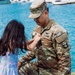 A U.S. Army soldier named TIRO, wearing Operational Camouflage Pattern (OCP) uniform, kneels down as a young girl pins or adjusts something on his chest near a waterfront. The scene appears to be a military ceremony, likely a promotion or homecoming event, suggested by the presence of a purple orchid lei being held nearby. The setting appears to be in Hawaii, indicated by the traditional lei and the marina visible in the background.