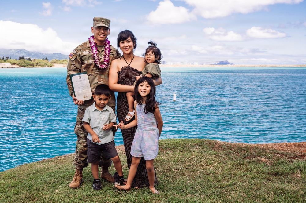 A U.S. Army soldier in camouflage uniform wearing a purple lei poses with his family near a body of turquoise water, likely in Hawaii. He is holding an official military certificate or document, suggesting a promotion, award, or recognition ceremony. The group includes a woman and three young children, all smiling for the photo.
