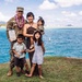 A U.S. Army soldier in camouflage uniform wearing a purple lei poses with his family near a body of turquoise water, likely in Hawaii. He is holding an official military certificate or document, suggesting a promotion, award, or recognition ceremony. The group includes a woman and three young children, all smiling for the photo.