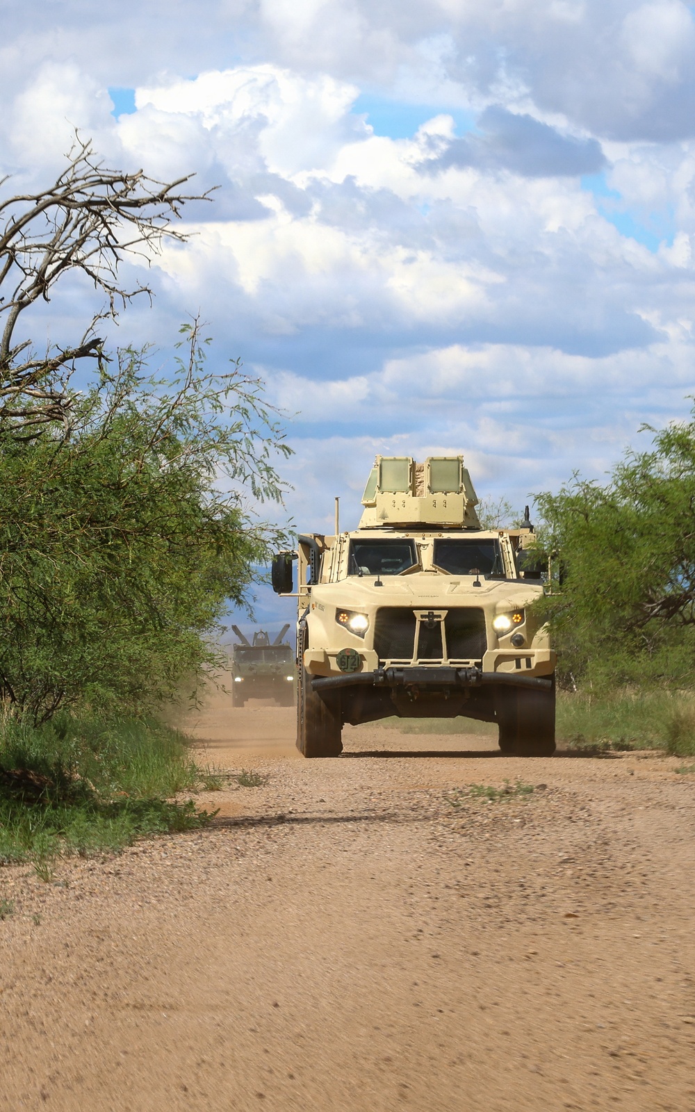 A convoy of military tactical vehicles travels along a dusty dirt road through a semi-arid landscape with scrub vegetation. The lead vehicle is a tan/desert-colored Joint Light Tactical Vehicle (JLTV) with a roof-mounted weapons system, followed by at least one additional military vehicle visible in the background. The scene appears to be a training or patrol operation in an arid environment, likely in the American Southwest based on the vegetation and terrain.