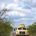 A convoy of military tactical vehicles travels along a dusty dirt road through a semi-arid landscape with scrub vegetation. The lead vehicle is a tan/desert-colored Joint Light Tactical Vehicle (JLTV) with a roof-mounted weapons system, followed by at least one additional military vehicle visible in the background. The scene appears to be a training or patrol operation in an arid environment, likely in the American Southwest based on the vegetation and terrain.