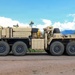 A large military heavy-duty recovery or wrecker vehicle is photographed in profile view on a gravel surface. The vehicle is painted in desert tan/sand camouflage and features multiple axles, heavy-duty equipment, and what appears to be a crane or recovery arm. The background shows a semi-arid landscape with mountains, vegetation, and a partly cloudy blue sky.