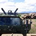 The image shows a military vehicle compound in an arid, semi-desert environment with mountains visible in the background. A dark green military truck in the foreground bears USMC markings and is equipped with what appears to be a dual-barrel weapon system or launcher mounted on top. Several additional tan/desert-camouflage military vehicles are visible in the background near a small structure.