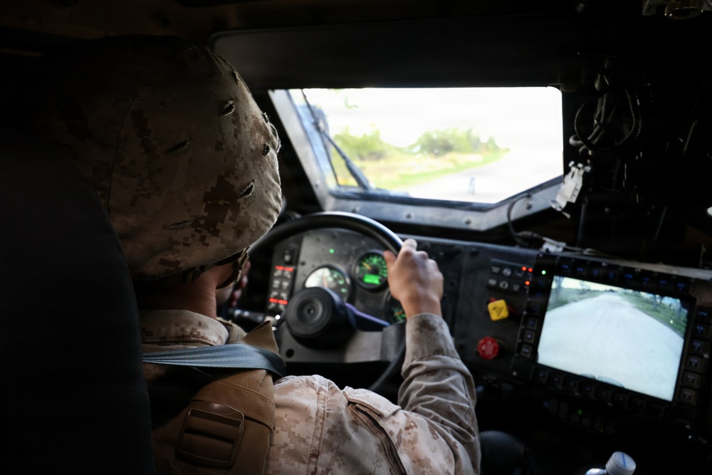 A military personnel member in desert camouflage uniform and helmet is driving a military tactical vehicle, likely a HMMWV (Humvee). The interior shows a complex dashboard with multiple controls, indicator lights, and a rear-view camera monitor. The vehicle appears to be in motion on a paved road based on the view through the windshield.