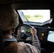 A military personnel member in desert camouflage uniform and helmet is driving a military tactical vehicle, likely a HMMWV (Humvee). The interior shows a complex dashboard with multiple controls, indicator lights, and a rear-view camera monitor. The vehicle appears to be in motion on a paved road based on the view through the windshield.