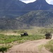 Two military vehicles are traveling along a dusty dirt road through an open grassland valley with rugged mountains in the background. The lead vehicle appears to be a heavy military truck with equipment mounted on it, while a flatbed truck follows behind, both kicking up dust clouds. The terrain suggests an arid or semi-arid environment, possibly a training area or remote military installation.