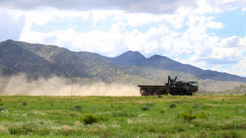 A large military truck is driving across an open field or dirt track, kicking up a significant dust cloud behind it. The vehicle is set against a backdrop of rugged mountain terrain with scattered vegetation. The scene appears to be a military training or operational exercise in an arid, semi-rural environment.