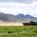 A large military truck is driving across an open field or dirt track, kicking up a significant dust cloud behind it. The vehicle is set against a backdrop of rugged mountain terrain with scattered vegetation. The scene appears to be a military training or operational exercise in an arid, semi-rural environment.
