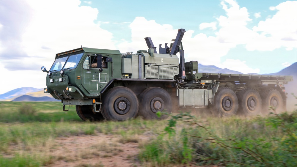 A large military heavy tactical truck, painted in olive drab green, is traveling at speed along a dirt road in an open field environment. The vehicle appears to be a multi-axle logistics or recovery truck, likely a variant of the HEMTT (Heavy Expanded Mobility Tactical Truck) or similar platform, equipped with a crane/loading arm mounted behind the cab. Dust is being kicked up from the rear wheels, and mountains are visible in the background under a partly cloudy sky.
